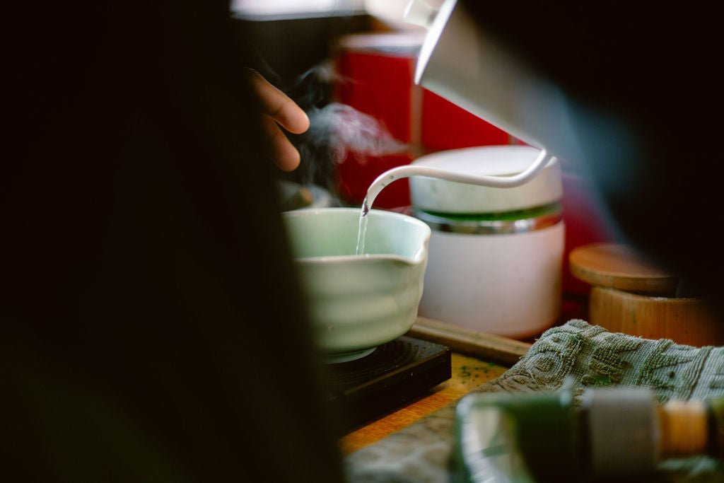 Hands pouring hot water over ceremonial grade matcha powder in a white bowl