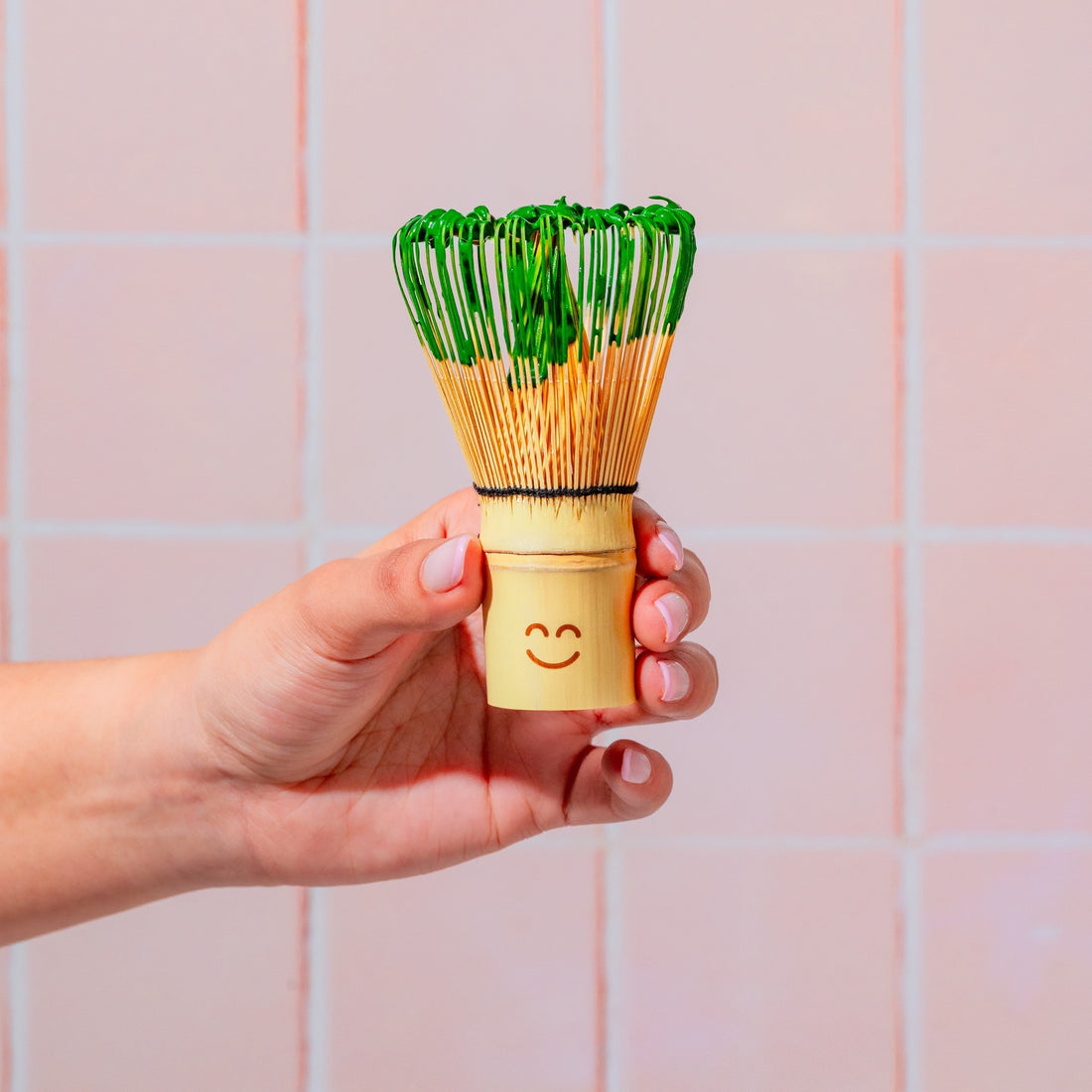 Hand holding a bamboo matcha whisk on a pink background